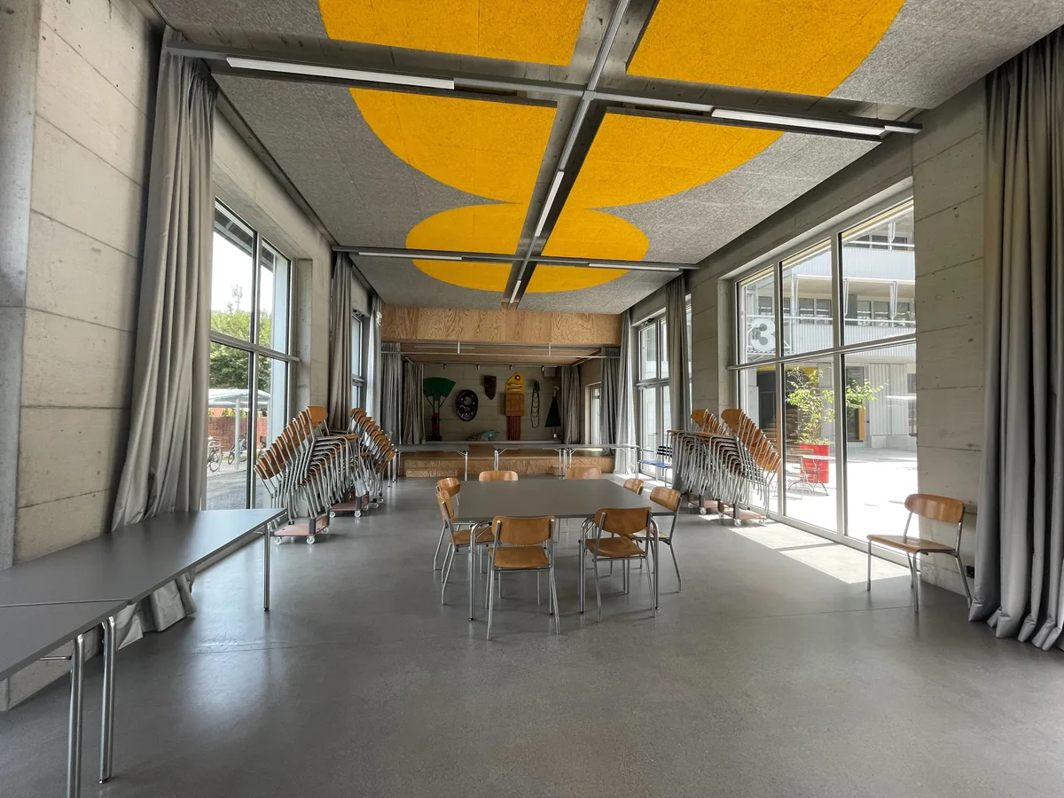 Der Mehrzwecksaal kann von Bewohnern des Westhofs benutzt und auch von Externen gemietet werden.  Bild einer Mehrzweckhalle mit Stühlen und Tischen und Kunst an Decke und Wänden.